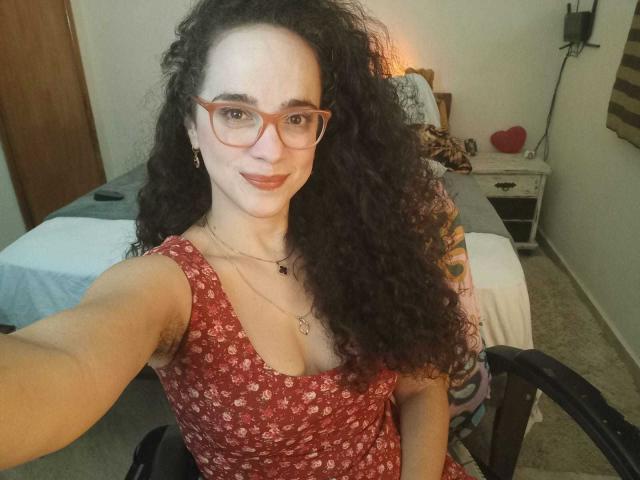 AmyFlores, met krullend zwart haar, rode bril en een rode jurk met bloemenprint, glimlacht naar de camera en laat haar kleine borsten en getatoeëerde arm zien, terwijl ze in een stoel zit..