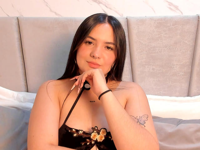 ValeriJoness with long black hair, light skin, small nose ring, black lace top, showing tits, arm tattoo of butterfly, resting chin on hand, smiling gently, bed in background.