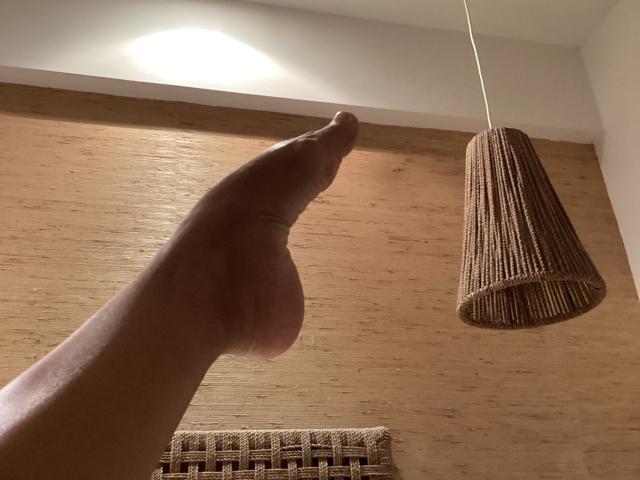 DeesseBleue's hand is pointing at a brown, woven, cone-shaped lamp hanging from a white cord. The lamp's texture is ribbed and it's suspended in a warmly lit room.