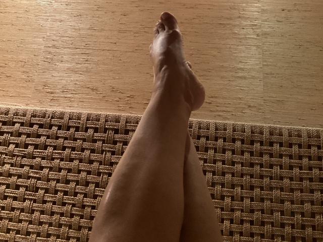 DeesseBleue's legs are crossed at the ankles, showing her bare, smooth, and slightly tanned feet with a relaxed, upward posture against a woven, brown fabric.