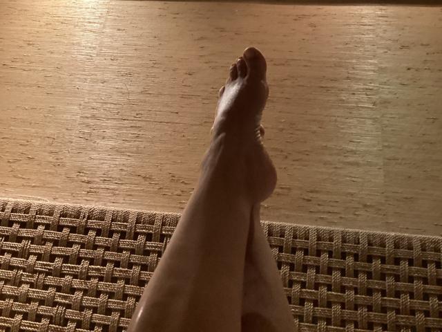 DeesseBleue's bare legs with feet crossed at the ankles, showing her toes, resting on a woven, beige, textured surface under warm, ambient lighting.