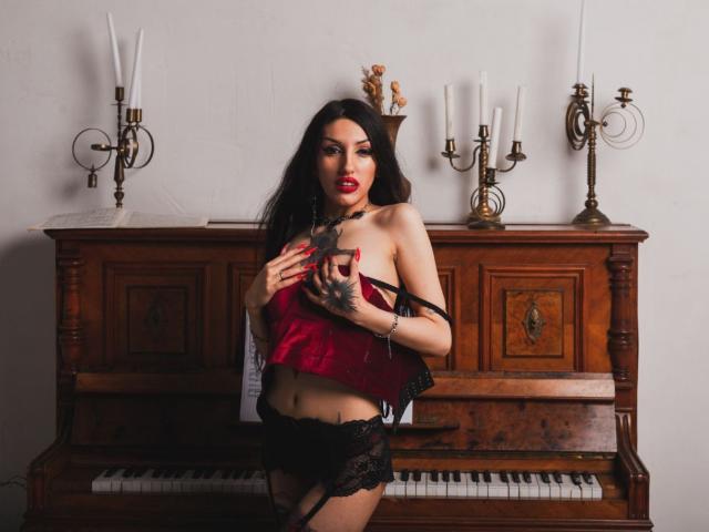StellaDarkspace with red satin bustier, black lace panties, and garter, holding her tits, standing in front of a wooden piano, red lipstick, long black hair, and tattoo on hand.