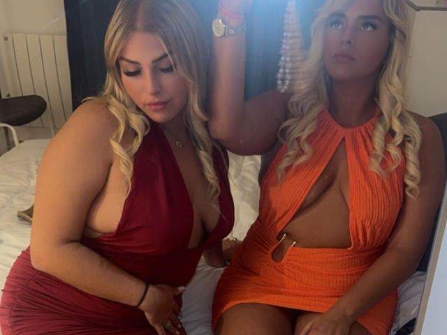 KenzaX69 with blonde hair, deep red dress, showing her tits, next to KenzaX69 in an orange dress, showing her tits, sitting on a bed, taking a selfie.