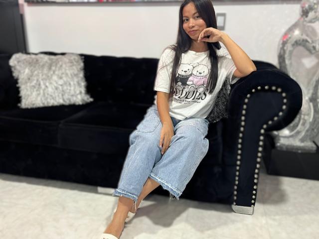 MilaBennet sitting on a black velvet couch, wearing a white "Snoo" tee, blue jeans, and white heels, with long dark hair, smiling, and her hand on her chin.