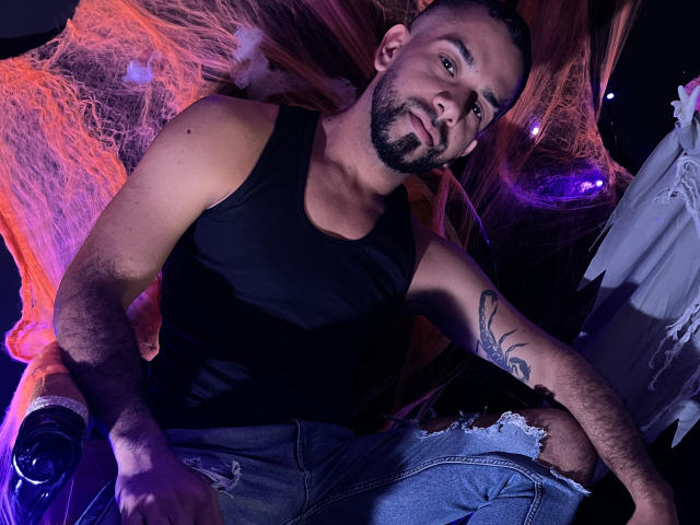 LoboSexS, un uomo muscoloso con una canotta nera, jeans strappati e tatuaggi, è appoggiato a delle cinghie arancioni con un'espressione sicura e sensuale. Sta tenendo una bottiglia..