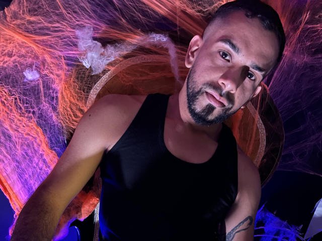 LoboSexS, un uomo muscoloso con una barba curata e una canotta nera, ha un'espressione seria e un tatuaggio sul braccio. Fumo arancione e viola lo circonda..