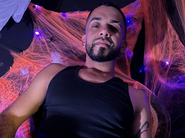 LoboSexS è un uomo muscoloso con una barba curata, una canotta nera e un tatuaggio sul braccio destro, sdraiato su una ragnatela arancione e viola, che sorride alla telecamera..