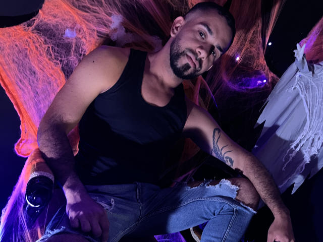 LoboSexS, con barba, canotta nera e jeans strappati, tiene in mano una bottiglia, ha tatuaggi sul braccio ed è circondato da luci arancioni e viola..