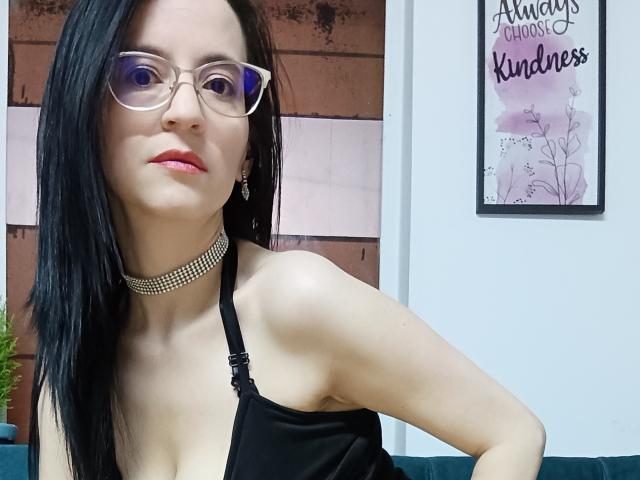 AlanaMoneth met zwart haar, een bril, roze lippenstift en een choker, draagt een zwarte haltertop, waardoor haar kleine borsten en lichte huid zichtbaar zijn. Ze leunt iets voorover..