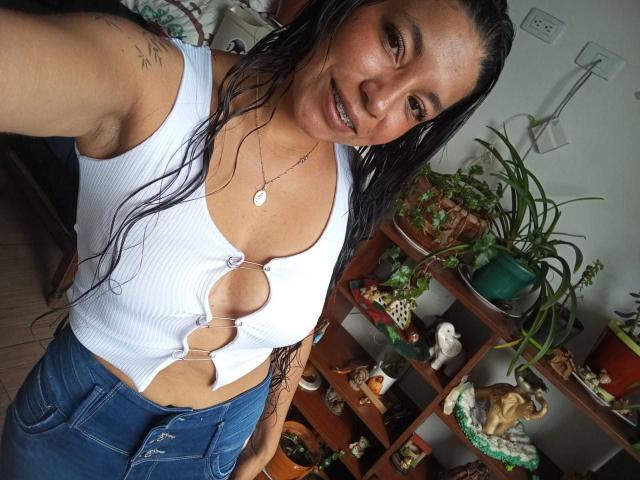 Paulina Colemann, con i capelli neri bagnati, sorride, indossa un top corto bianco a coste con aperture a cuore che rivelano il seno, e jeans blu, ed è in piedi in una stanza con delle piante..