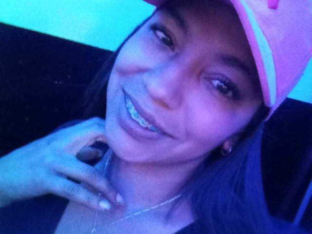 Paulina Colemann, avec une peau légèrement bleutée, portant une casquette rose, souriant avec des bagues orthodontiques, la main près du cou, de longs cheveux bruns, de petites boucles d'oreilles et un collier en argent..