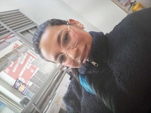 Paulina Colemann, con gafas, un cuello alto negro y suave, y reflejos azules en su cabello oscuro, sonríe ligeramente, mostrando su pequeño trasero. Su piel morena es lisa. Está en el interior, mirando a la cámara..