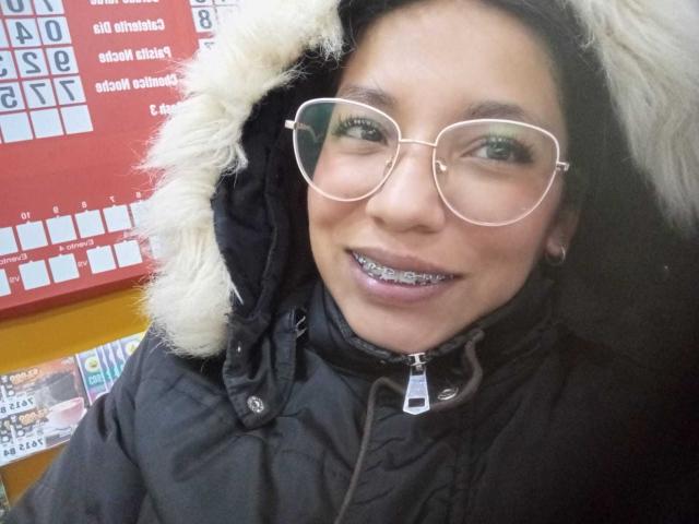 Paulina Colemann, usando óculos, com um casaco preto acolchoado, capuz de pele branca, sorrindo, com "grills" de metal nos dentes, pele clara, cabelo preto e olhos castanhos, em pé em frente a uma parede vermelha..