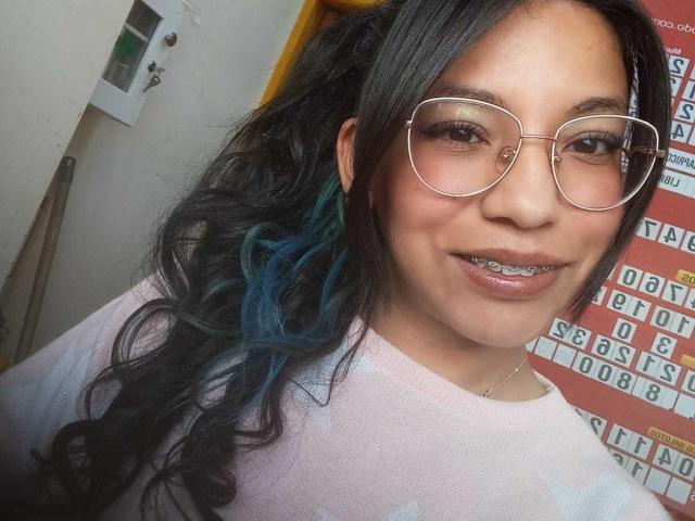 PaulinaColemann with black wavy hair, blue highlights, wearing glasses, pink sweater, and metal teeth, smiling at the camera. Her tits are small and she has a cute face.