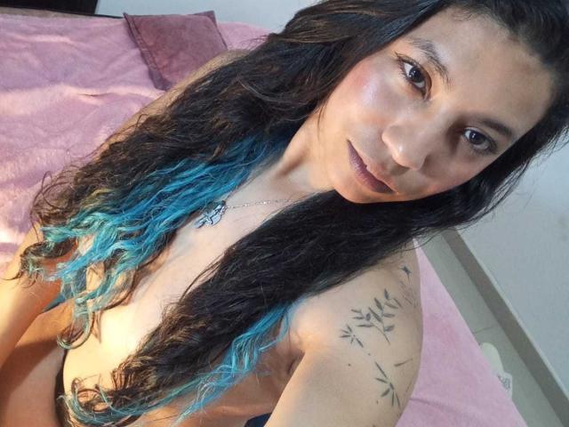 PaulinaColemann with long black and blue ombre hair, small tits, tattoo on right shoulder, sitting on pink bed, looking at camera with slight smile, wearing black top.