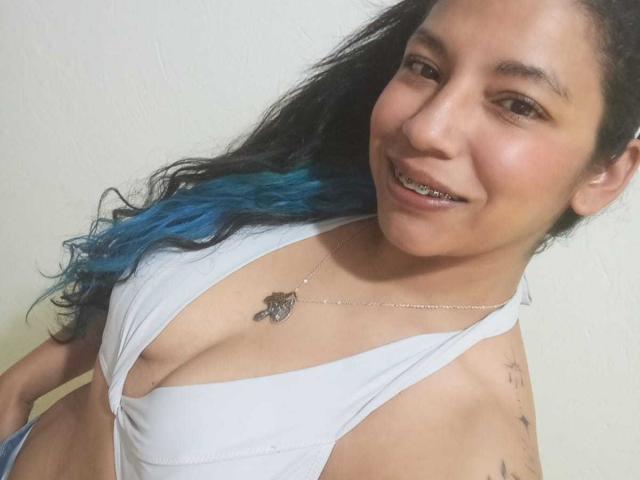 PaulinaColemann with blue-tipped hair, smiling, showing tits in a white halter top, silver necklace, and tattoos on her arm. She has a lip piercing and medium skin.