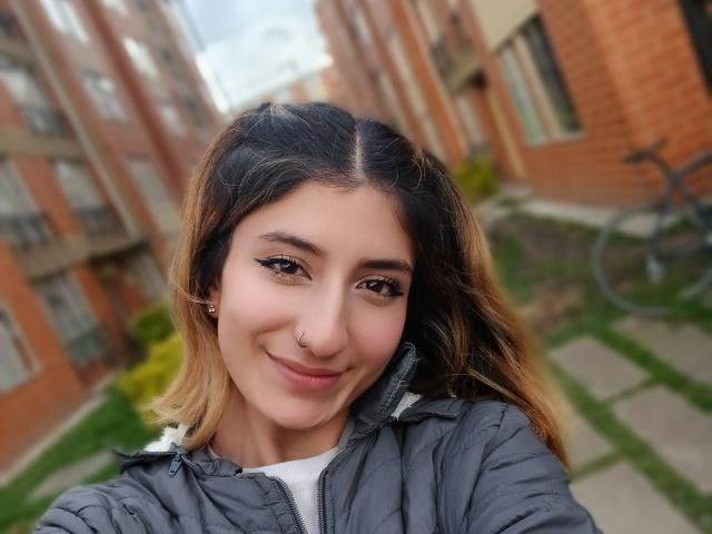 Kaiix met bruin en blond haar in een ombre-stijl, draagt een grijze, gewatteerde jas, glimlacht, heeft een neusring, zwarte eyeliner, een wit shirt en staat in een smalle, stedelijke binnenplaats met een fiets..