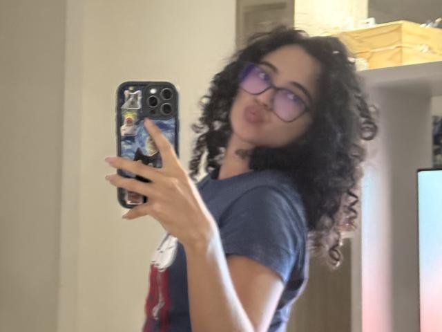 Vivienne Laurent, con cabello negro rizado, gafas moradas y una camiseta azul, hace pucheros frente al espejo mientras sostiene un teléfono con una funda de colores. No se ven los senos..