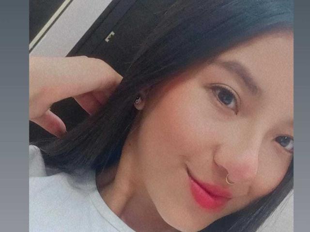 NaiBlue has dark hair, a nose ring, red lipstick, and a slight smile, with a hand in her hair, wearing a white shirt. Her face is close to the camera.