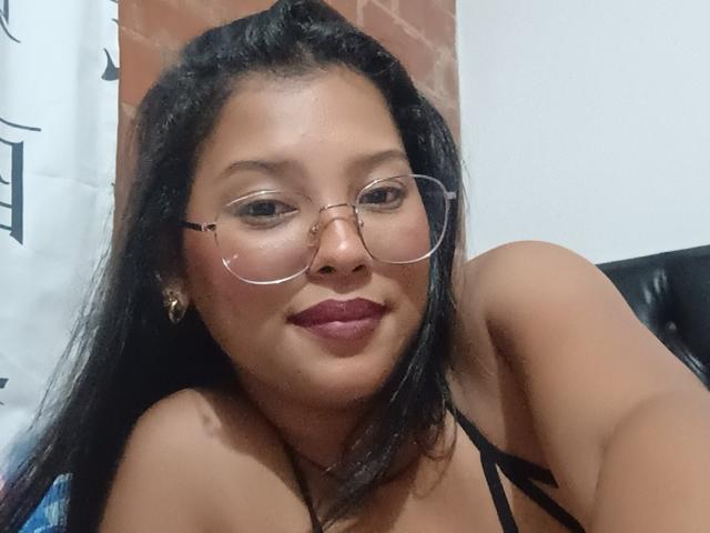 KolorBella has medium brown skin, long black hair, wears glasses, and has red lipstick. She's smiling, showing her tits in a black strappy top. Her ass is not visible.