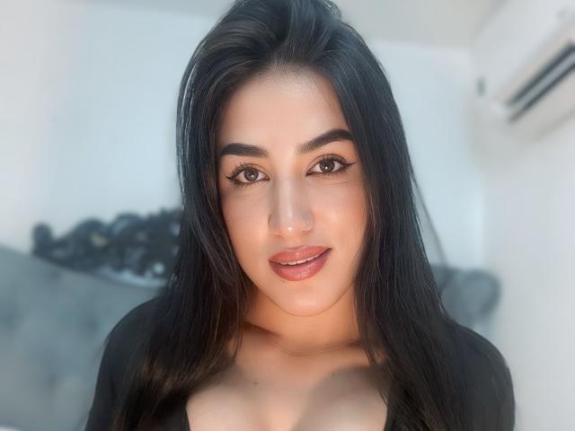 AlanaHarris has long black hair, brown eyes, and full pink lips. She's wearing a black top, showing her tits slightly. She's smiling subtly, looking directly at the camera.