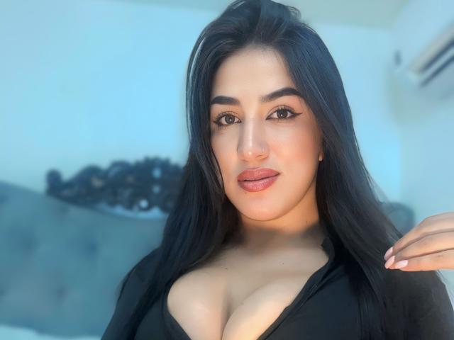 Alana Harris tem cabelos pretos longos, sobrancelhas grossas e lábios carnudos. Ela está vestindo uma blusa preta, que mostra seus seios grandes, e tem um tom de pele claro, olhando para a câmera..