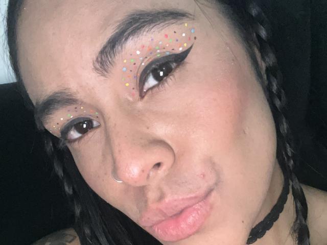 StellaBeckker has colorful glitter on her eyelids, black eyeliner, and is pouting her lips with a nose ring and black choker. She has braided hair and light brown skin.