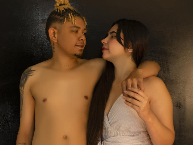 EmmaHarding en haar partner, zonder shirt en met een knot, met een tatoeage op zijn schouder, kijken elkaar aan. Ze draagt een witte jurk, heeft haar arm om zijn nek, donker haar en rode lippen..