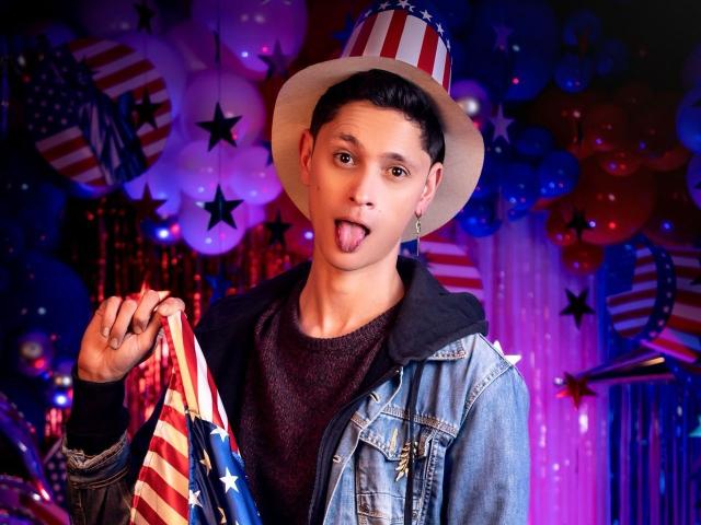 TylerCa with short black hair, wearing a white hat with red and blue stripes, denim jacket, and dark shirt, sticking out his tongue, holding a flag in his right hand.