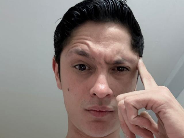 TylerCa has dark slicked-back hair, a small mole on his left cheek, and a serious expression while touching his forehead with his right hand. His brown eyes are slightly squinted.