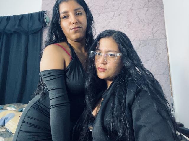 AdelaandSharick with long black hair, wearing a black dress and red bra strap, standing next to AdelaandSharick in glasses, black jacket, and black hair, looking at camera.