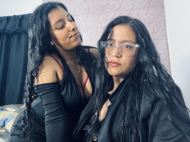 AdelaandSharick with long black hair, wearing a black dress and red bra, leaning on AdelaandSharick in glasses, black shirt, and black hair, both with serious expressions.