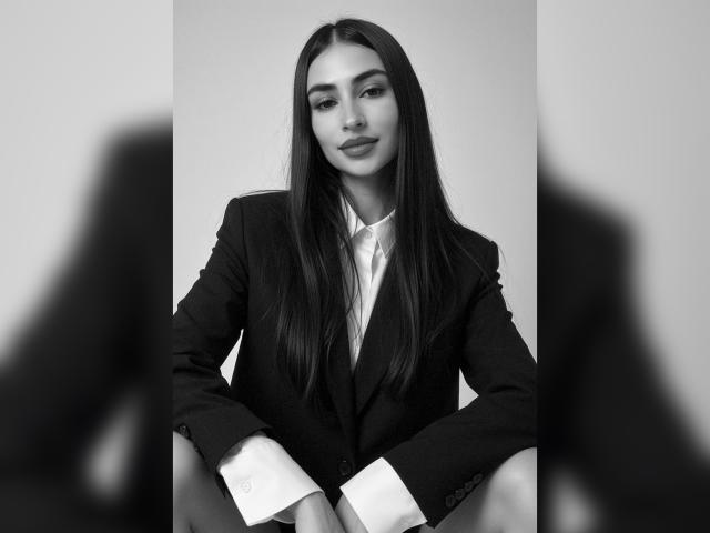 Foto em preto e branco de MiilaWx com cabelo longo e liso e escuro, vestindo um blazer escuro sobre uma camisa branca, sentada com um leve sorriso, mostrando seus seios lisos..