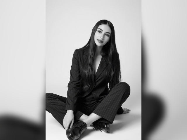 MiilaWx with long black hair, sitting cross-legged, wearing a pinstriped suit, buttoned-up, and shiny black shoes, looking at the camera with a slight smile. Black and white photo.