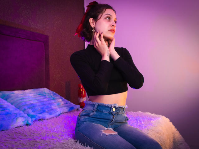 Sophie Blake, con le mani sul mento, indossa un top corto nero, jeans blu strappati e un fiocco rosso tra i capelli, seduta su un letto con illuminazione viola e blu..