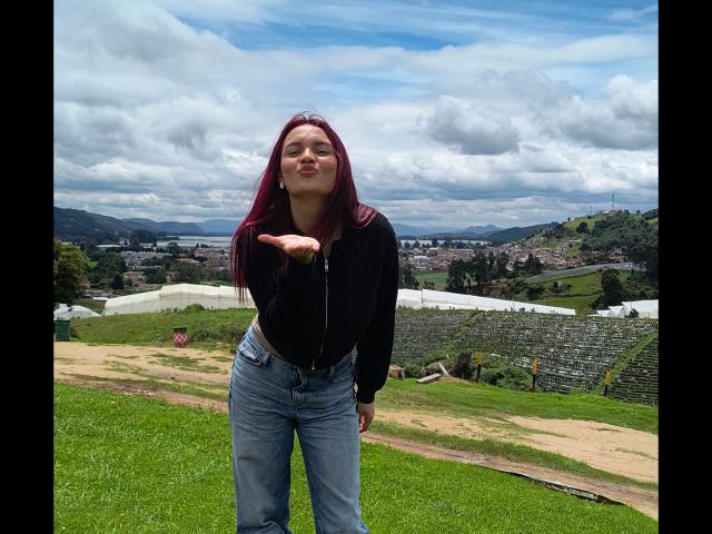 JulietaFoster69 with red hair, black top, and blue jeans, smiling, pointing forward, standing on grassy hill with stone terraces and cloudy sky in the background.