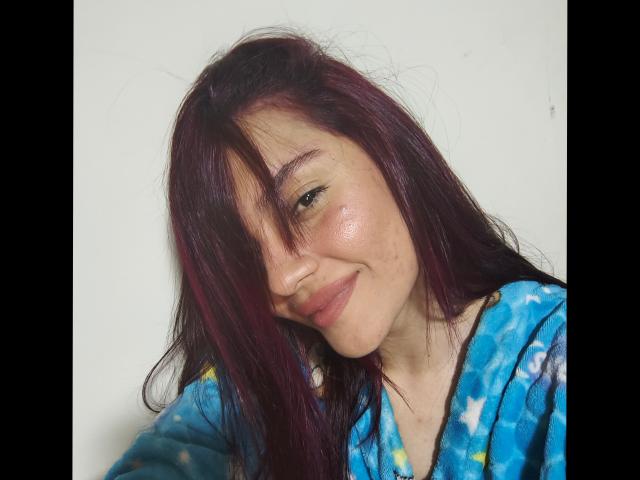JulietaFoster69 with dirty purple hair, smiling slightly, wearing a blue tie-dye shirt, has a cute face and shiny skin, looking to the side with a relaxed expression.