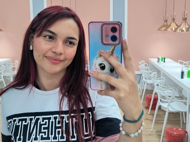 JulietaFoster69 with red hair, white shirt, holding pink phone with unicorn case, smiling, white earbuds, pink wall, white chairs, and pink table in the background.