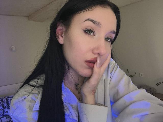 NikkiGoldX has pale skin, black hair, and is resting her face on her hand, wearing a white hoodie with a blue pattern, looking at the camera with a slight smile.