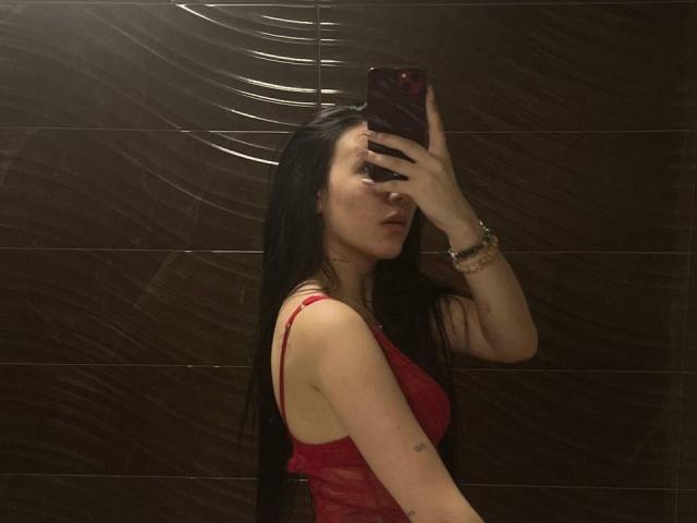 NikkiGoldX with long black hair, red lace top, holding phone in mirror selfie, showing side boob, wrist bracelet, subtle tattoo on arm, dark tiled wall background.