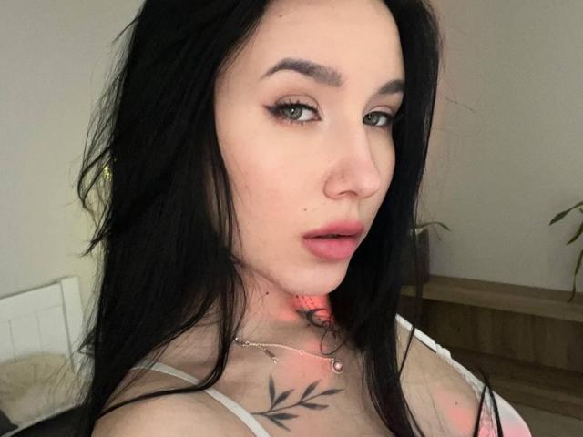 NikkiGoldX has long black hair, green eyes, and a subtle tattoo under her chin. She's wearing a white top with a pink necklace, looking at the camera.
