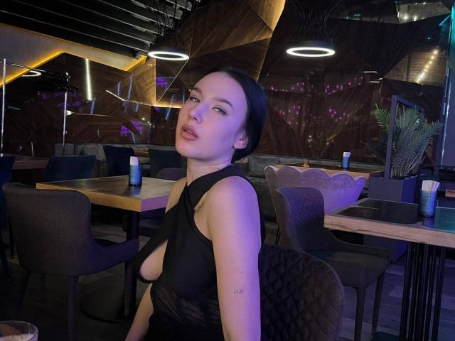 NikkiGoldX met kort zwart haar, bleke huid en kleine tatoeages, draagt een zwarte, laag uitgesneden, doorschijnende top die haar borsten laat zien, in een zwak verlicht restaurant, en kijkt naar de camera..