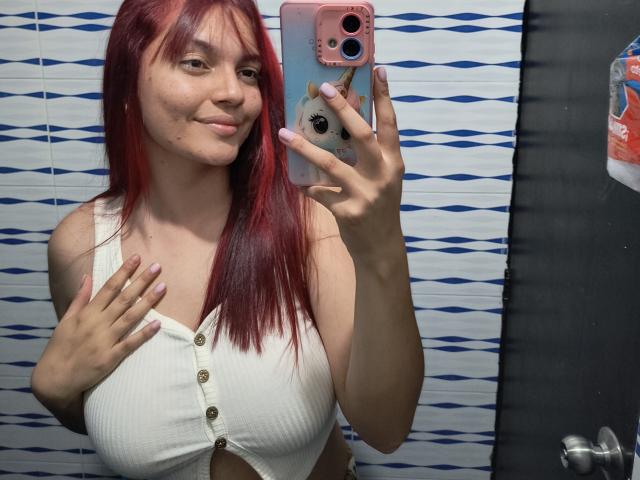 JulietaFoster69 with red hair, smiling, holding phone, wearing white ribbed crop top with buttons, showing her boobs, pink nails, in bathroom mirror selfie.