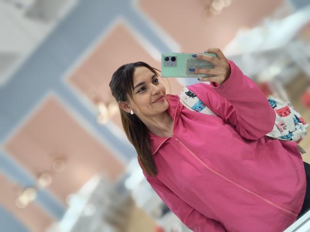 JulietaFoster69 with brown hair in a ponytail, pink jacket, white floral bag, holding green phone, smiling at mirror, pink earrings, blue nail polish, indoors, selfie.