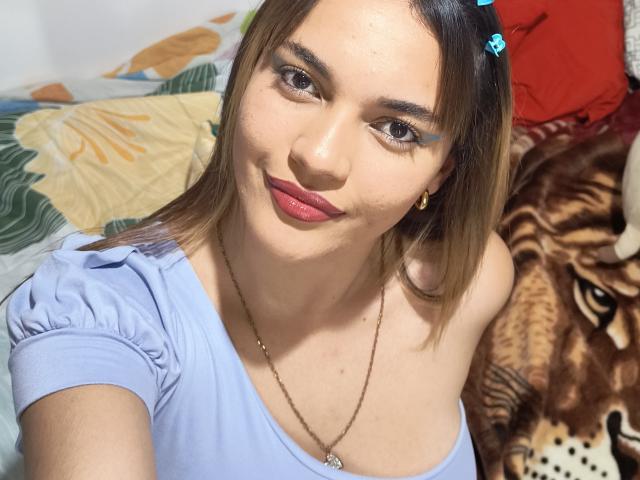 JulietaFoster69 with light skin, blue eyes, and brown hair, wearing a blue top, blue eyeliner, pink lipstick, gold earrings, and a necklace, smiling at the camera.