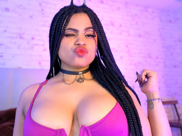 AprilKershaw with long black braids, pink bra, big tits, choker, red lipstick, winking, holding nail file, pink and purple lighting, smiling.