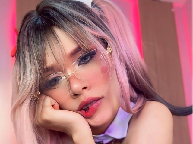 ZoeJonesOne with pink and blonde pigtails, wearing glasses, and pink lipstick, resting her chin on her hand, looking sexy with red blush on her cheeks.