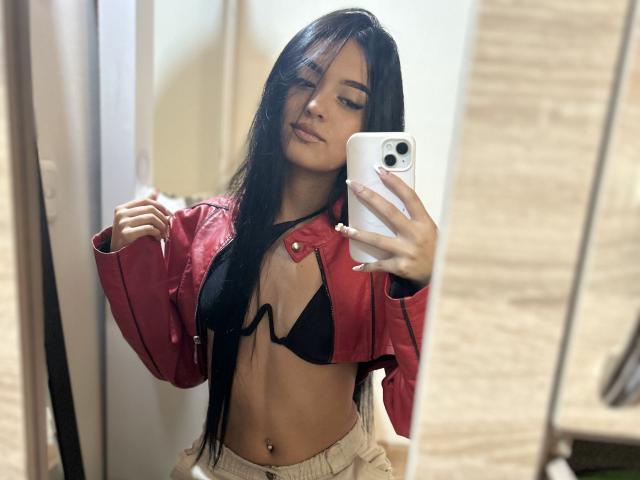 AmySaens with long black hair, red jacket, black bra, and beige pants, taking a mirror selfie, showing her small tits and belly button, holding a white phone.
