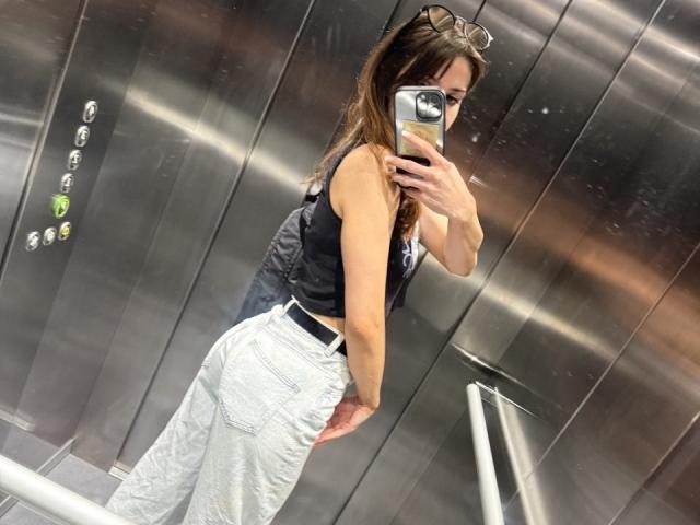 SweetyFit with long brown hair, wearing a black tank top and white high-waisted jeans, taking a selfie in a silver elevator, showing her ass.