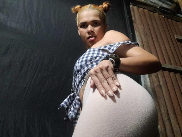 DomiEvans, avec un chignon orange, portant une chemise bleue à carreaux avec une épaule dénudée, un pantalon blanc moulant, une main sur ses fesses, regardant vers le bas, un bracelet noir, des ongles décorés, et une expression confiante..